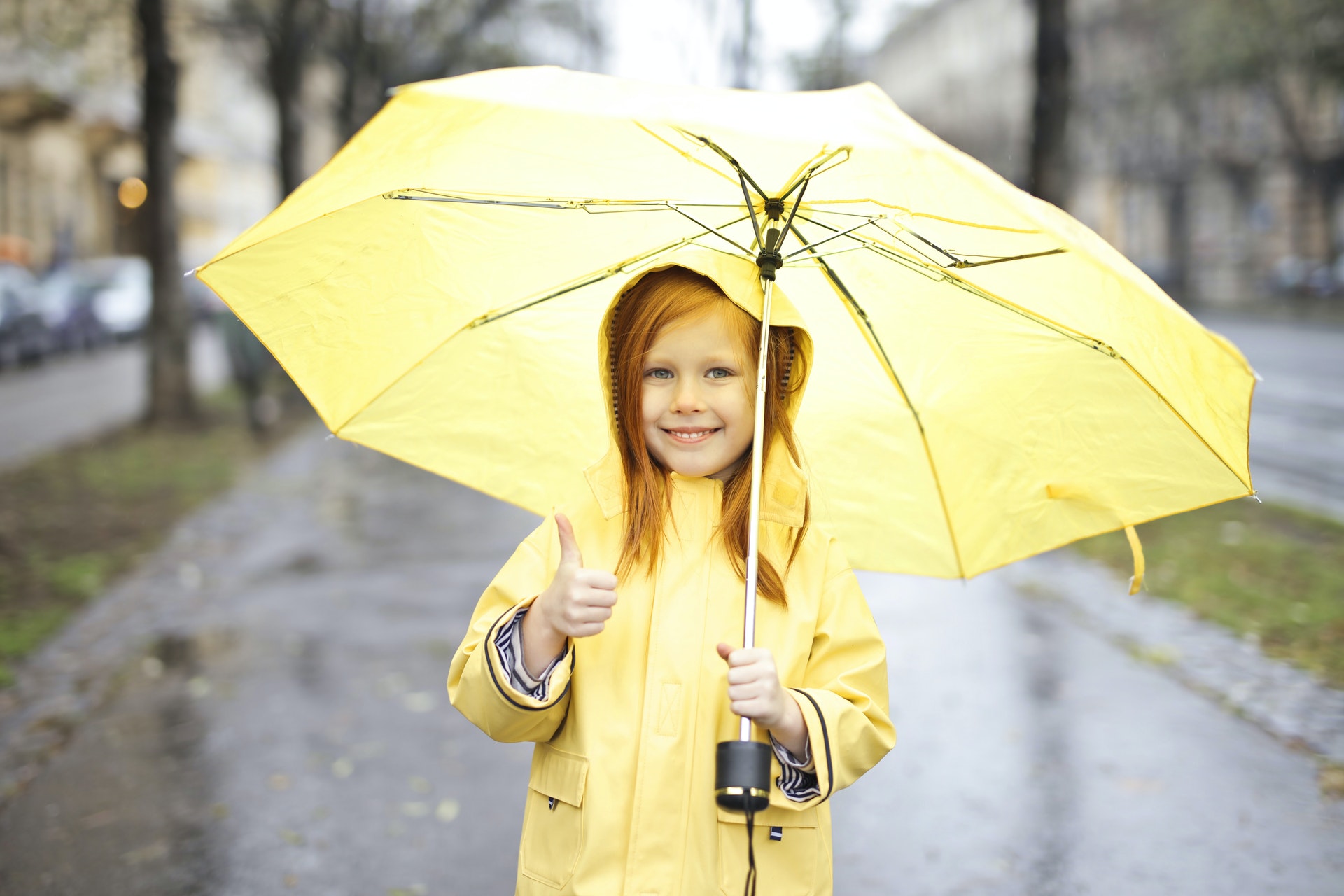 Photo of Smiling Girl in Yellow Raincoat Holding a Yellow Umbrella While Giving One Thumbs Up