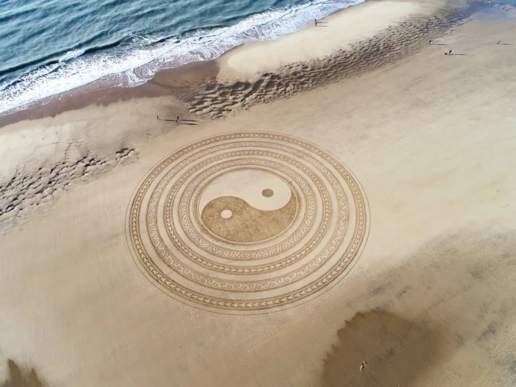 A yin yang symbol cut into beach sand that's about to be hit by a wave.