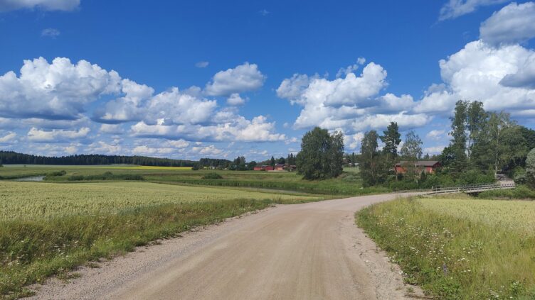 Dirt road on the right, green field on the left, river passing below bridge and clear blue sky.
