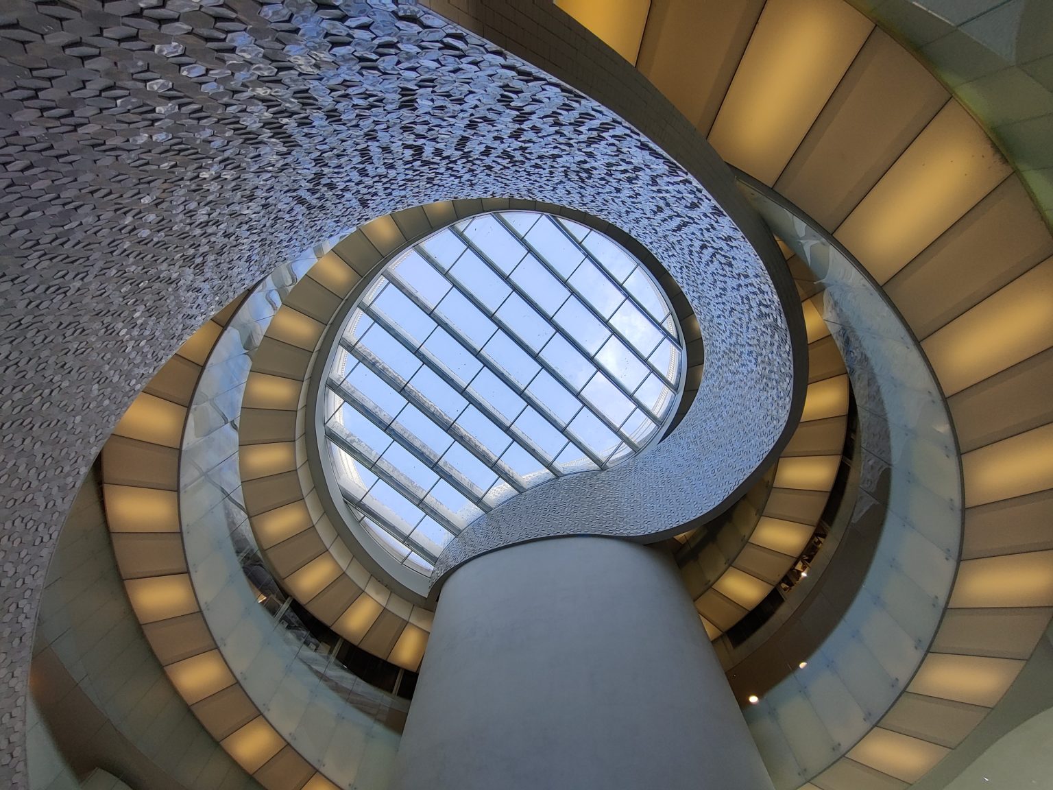 An interior architectural detail featuring a circular design. There's a textured, patterned ceiling leading to a round skylight. Adjacent to this is a spiral structure with illuminated segments. A pillar supports the structures. The overall color palette is white and beige.