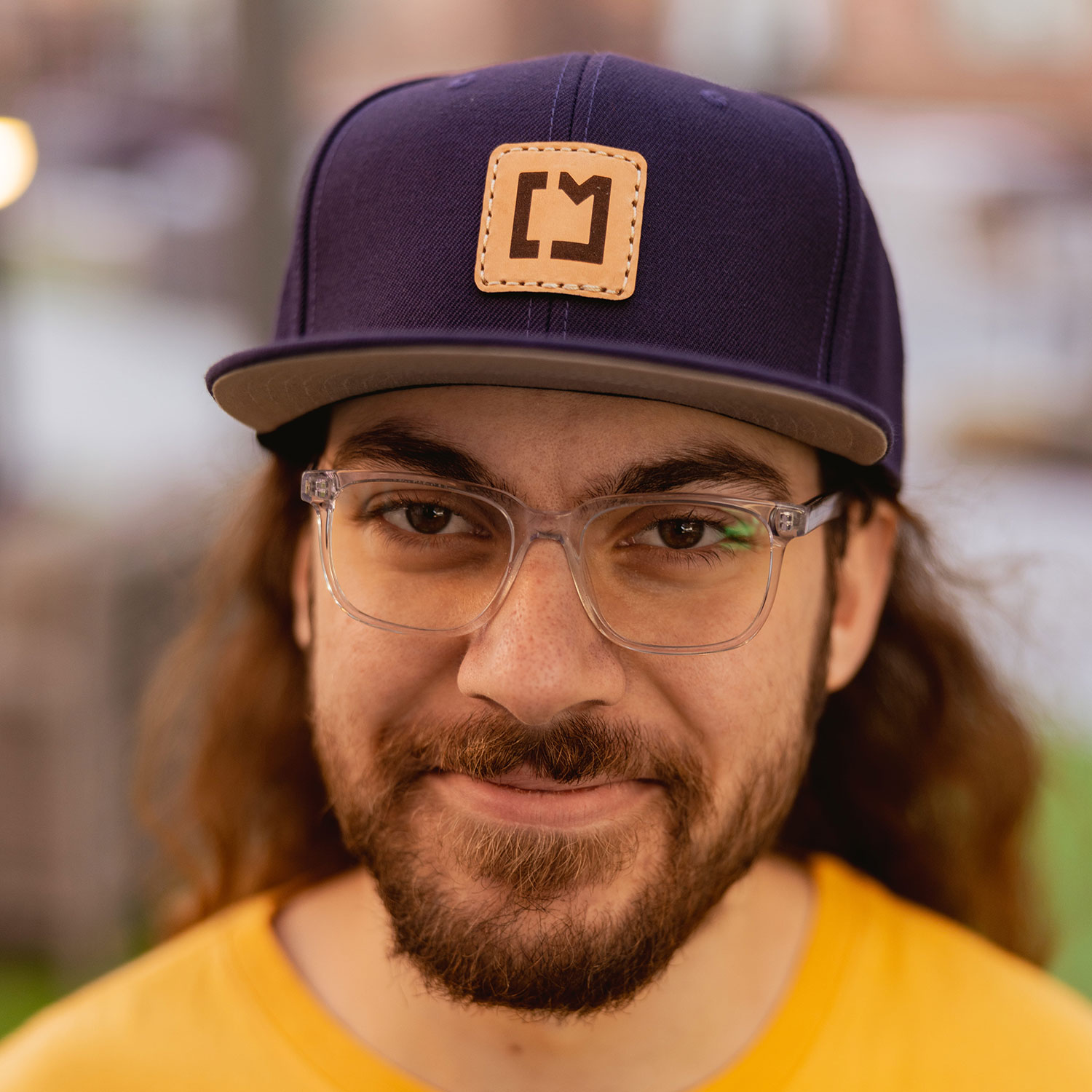 Headshot: Christian Taylor. He has a beard and long hair. He is wearing a yellow t-shirt, glasses, and a dark baseball cap with his logo on it, a stylized "CM"